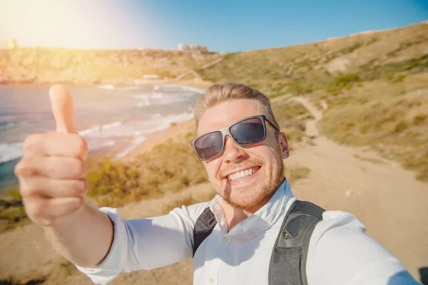Erkek gezgin arka planda mavi deniz, kumsalda güneş gözlüklü ve sırt çantalı selfie çekiyor. Seyahat kavramı — Stok fotoğraf