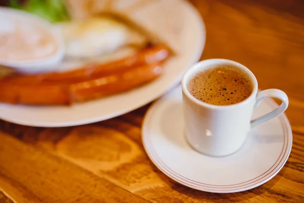 Klassisches englisches Frühstück mit Espressokaffee im weißen Becher, zwei Würstchen, Spiegeleiern, Toasts, Salat auf Holzgrund — Stockfoto