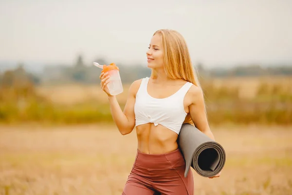 Güzel sarışın kız parkta halı üzerinde spor yogası yaptıktan sonra su içiyor. Egzersiz konsepti — Stok fotoğraf