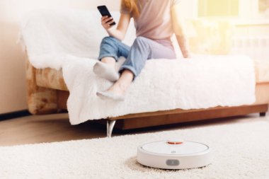 Halıda çalışan akıllı robot elektrikli süpürge kullanan genç bir kız. Telefonlu kontrol aygıtı