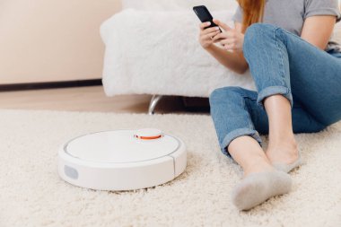 Halıda çalışan akıllı robot elektrikli süpürge kullanan genç bir kız. Telefonlu kontrol aygıtı