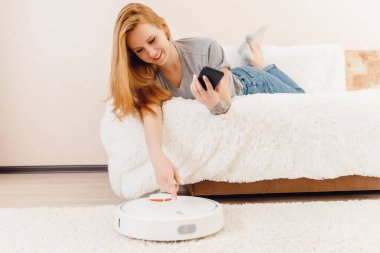 Halıda çalışan akıllı robot elektrikli süpürge kullanan genç bir kız. Telefonlu kontrol aygıtı