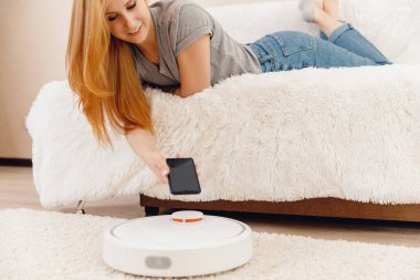 Halıda çalışan otomatik robot elektrikli süpürge kullanan genç bir kız. Akıllı ev fonksiyonlarını telefonla kontrol etmek