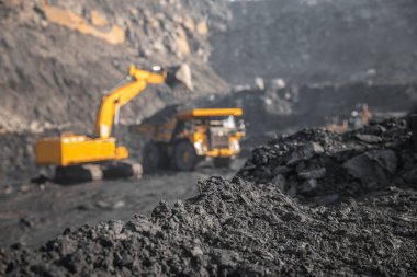 Sert siyah kömüre odaklan, endüstriyel taş ocağı benimkini aç. Arka plandaki bulanık mineral antrasit kazıcısını büyük sarı kamyona yükle