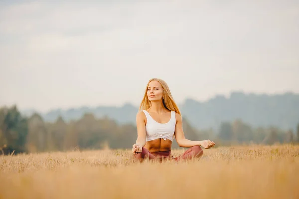Chica joven en camisa blanca, ropa deportiva con pelo largo Rubia medita yoga en campo amanecer. Concepto de relajación —  Fotos de Stock