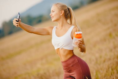 Güzel sarışın kız spor eğitimi sırasında telefonda fotoğraf çekiyor, gülümsüyor.
