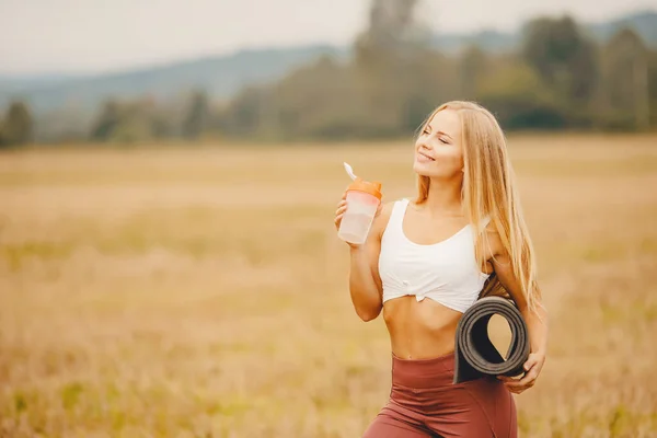 Belle fille blonde étanche et boit soif avec bouteille d'eau, tapis de yoga, dans le parc de fond — Photo