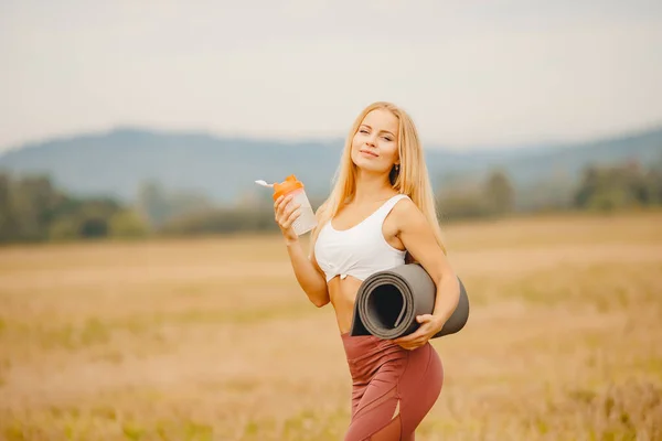 Belle fille blonde tient bouteille d'eau et tapis pour l'étirement et le yoga dans le parc — Photo