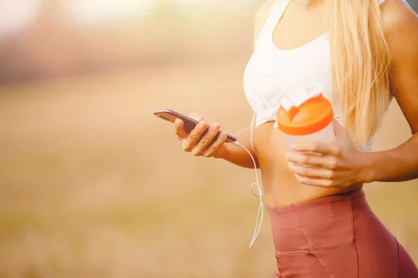 Menina usa telefone para treinamento on-line com fones de ouvido conectados, na garrafa de esportes de mão para água — Fotografia de Stock