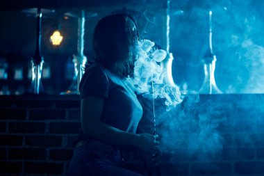 Güzel bir kadın gece kulübünde, nargile ya da shisha barında, mavi arka planda duman halkaları üfler.