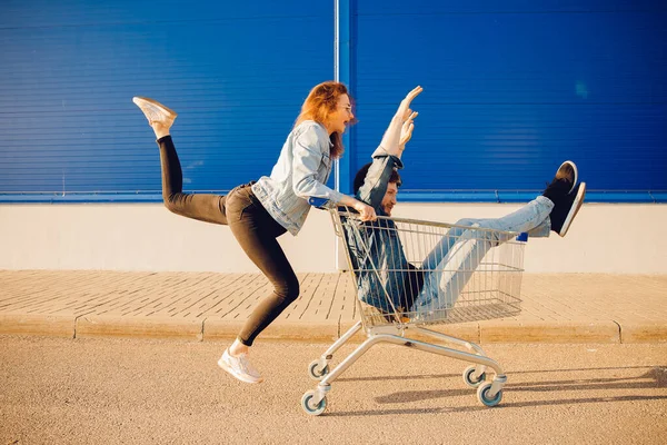 Supermarché, heureuse jeune femme portant l'homme dans le magasin de chariot pour le shopping, les mains levées. Couple famille coucher de soleil — Photo