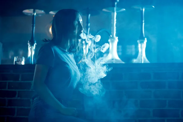 Молода красива жінка в нічному клубі курить кальян або шеша-бар, синій фон — стокове фото