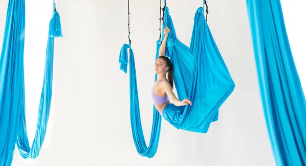 Ung flicka gymnast i sportkläder som sträcker sig på blå hängmatta i vit studio. Begreppet flygande yoga — Stockfoto