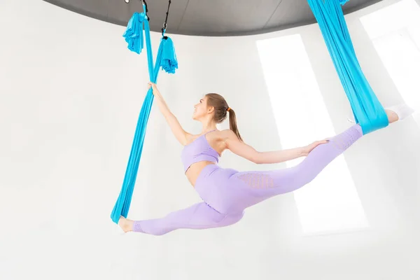Ung flicka gymnast i sportkläder som sträcker sig på blå hängmatta i vit studio. Begreppet flygande yoga — Stockfoto