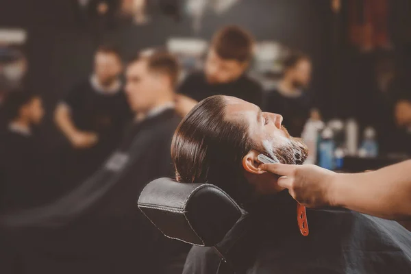Хипстер, сидящий в кресле парикмахера парикмахерская, бреет бороду опасной бритвой — стоковое фото