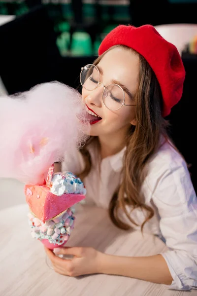 Wanita muda dengan baret merah dan kacamata memiliki makanan penutup manis besar dengan permen kapas merah muda di kafe di Perancis — Stok Foto