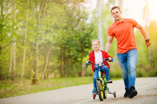 Pai ajuda seu filho a andar de bicicleta no ensolarado parque de dia — Fotografia de Stock