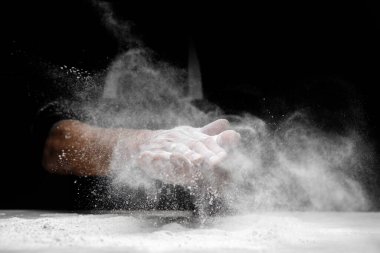 Beyaz toz un havaya püskürtülürken insan eliyle pişiren kişi siyah arka planda yemek pişiriyor.