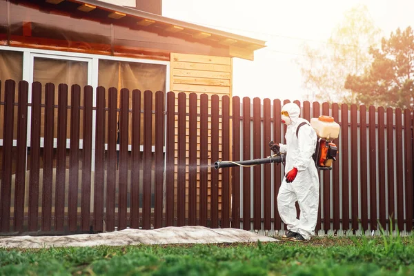 El desinfectador en el traje protector procesa el territorio de la parcela de jardín de la casa rocía el veneno de los mosquitos, las garrapatas y las plagas —  Fotos de Stock