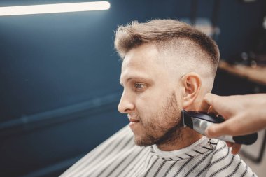 Berber dükkanında saç örgüsü yapan adama sakal kırpıyor. Elektrikli tıraş makinesi, renkli kahverengi.