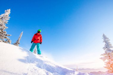 Arkaplanda masmavi bir gökyüzü ve Freeride kış ormanıyla uçan snowboardcu.