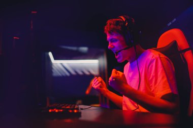 Oyuncu genç adam çevrimiçi bilgisayar oyunu oynuyor ve monitör turnuvayı kazanıyor, seviniyor ve el kaldırıyor, neon rengi