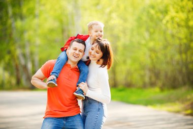 Oğullarını omuzlarında taşıyan ebeveynler yaz güneşi parkında yürürler. Çocuk gülümser ve mutlu anne ve baba kucaklaşır.