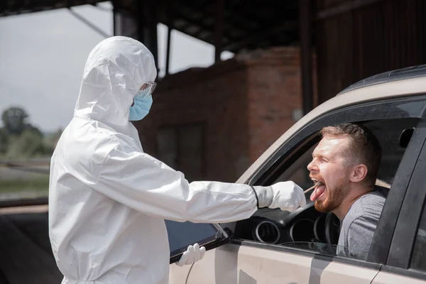 Concepto prohibición conductor coche viaje coronavirus. Médico en traje protector del virus que toma hisopo del hombre para evaluar la infección por COVID-19 — Foto de Stock