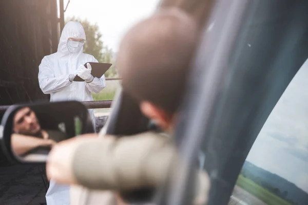 Ellenőrzöm az utasokat és a sofőrt, hogy van-e vírus, a biológiai öltönyös férfi adatokat ír táblagépre, autószámra és utazási útvonalra. fogalom Élet a világjárvány után — Stock Fotó