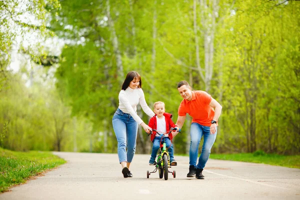 Pai e mãe ensina o pequeno filho a andar de bicicleta no parque, se divertir família — Fotografia de Stock