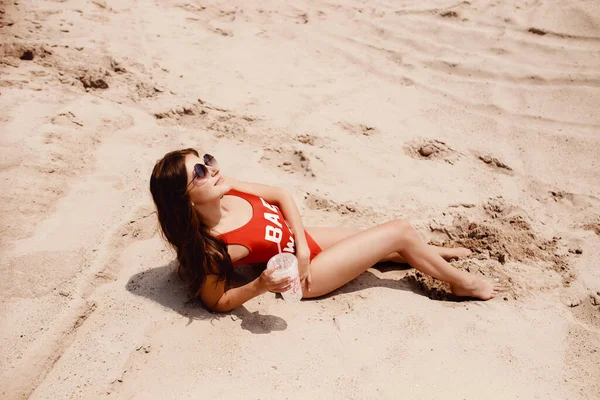 Relajante en la playa, la joven se encuentra en la arena en traje de baño rojo con una copa de cóctel y gafas de sol — Foto de Stock