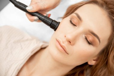 Doktor kozmetik spa prosedürü uyguluyor. RF, genç bir kadının cildine yapılan elektrikli yüz masajını kaldırıyor.