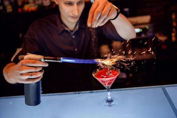 Бармен готовит красный коктейль с розмарином из корицы в прозрачном стекле на барной стойке с алкоголем. Использует одноразовые искры. Темный фон — стоковое фото