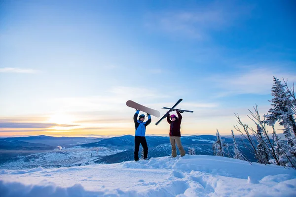 İki aktif dost snowboardcu ve kayakçı dağın tepesinde mavi gökyüzünde gün doğumunda duruyorlar. Kavramsal kayak merkezi kış ormanı — Stok fotoğraf