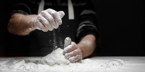 Baker chef hand sprinkling flour fresh dough on kitchen table, black background banner — Stock Photo, Image