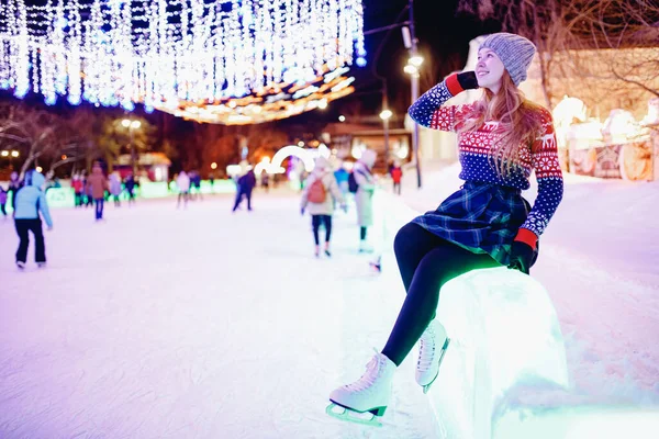 Beautiful young girl in warm red sweater on skating rink in figured white skates in evening against background of illumination — Stock Photo, Image