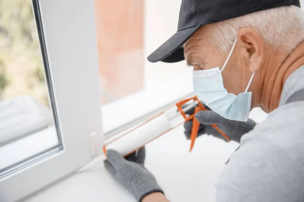 Trabalhador de construção em máscara médica usar tubo de silicone arma para reparar e instalar janela em casa — Fotografia de Stock