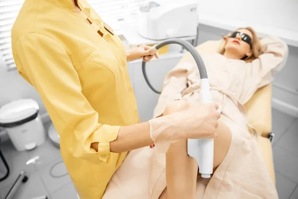 Косметик удаляет волосы медицинским лазером с ног красивой женщины в салоне красоты, белый фон — стоковое фото
