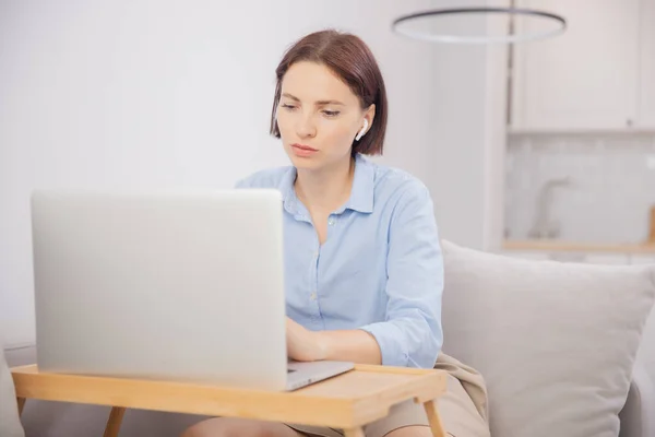 Lifestyle thuiskantoor, jonge vrouw in hoofdtelefoon werkt laptop via internet videogesprek vanaf de bank, lichte achtergrond — Stockfoto