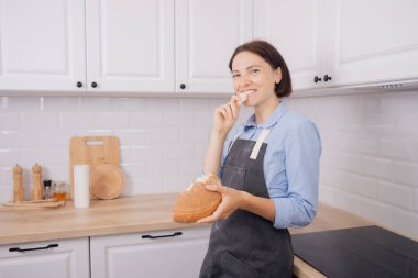 Genç kadın fırıncı beyaz mutfak arka planında taze pişmiş buğday ekmeği yiyor.