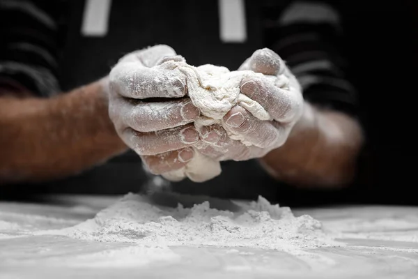 La farine blanche vole dans l'air sur fond noir, le pâtissier applaudit les mains et prépare la pâte à levure pour les pâtes à pizza — Photo