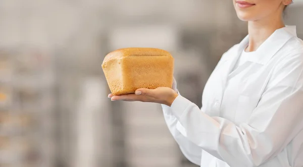 Munkás pék nő kezében kenyér a pékségben, háttér a gyártósor — Stock Fotó