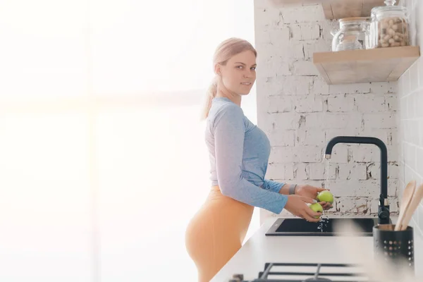 Concept healthy post workout nutrition, vitamin replenishment. Female in sportswear washes green apples in kitchen sink