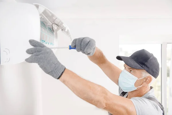 Técnico homem em máscara médica trabalhador de reparação e instala ar condicionado na parede branca — Fotografia de Stock