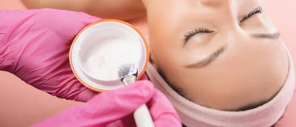 Lekarz stosuje krem przeciwzmarszczkowy starzenie skóry młodej kobiety w salonie piękności, różowe tło — Zdjęcie stockowe