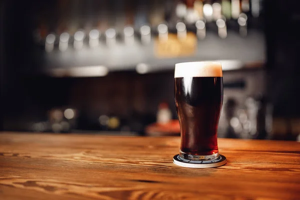 Bicchiere di birra scura artigianale con schiuma sul bancone del bar in legno, pub sfondo sfocato — Foto Stock