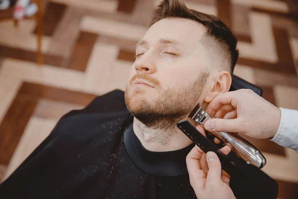 Крупный план парикмахера стригущей бороду мужчине в парикмахерской электрической бритвы — стоковое фото