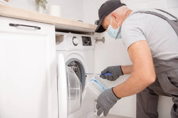 Working man in medical mask plumber repairs washing machine in kitchen