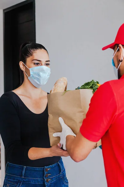 Mexican deliver man wearing face mask in red uniform handling bag of food, fruit, vegetable give to female costumer in front of the house. Grocery delivery service during covid19