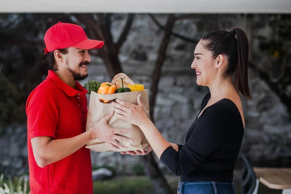 Latin deliver man in red uniform handling bag of food, fruit, vegetable give to female costumer in front of the house. Postman grocery delivery service in Mexico